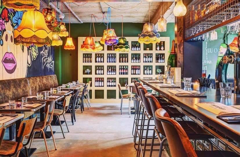 Salle d'I Love Burger à Visé : bar, banquette, abat-jour colorés et mur de bouteilles